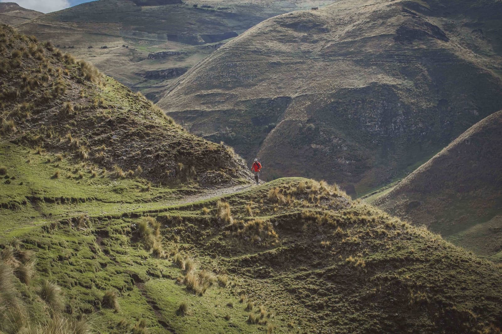 Um ponto vermelho no verde imenso dos Andes, e esse ponto é você. Um caminhante solitário com jaqueta vermelha percorre uma trilha estreita ao longo de um cume no Ancascocha Trek, cercado por colinas verdejantes cobertas de gramíneas andinas e vales profundos