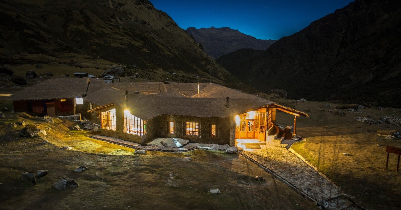 After a long day on the trail, this is exactly what you want to see. A stone-and-thatch mountain lodge glowing with warm amber light at dusk during an Andes trek, set in a high alpine valley with dark peaks rising under a deep blue twilight sky