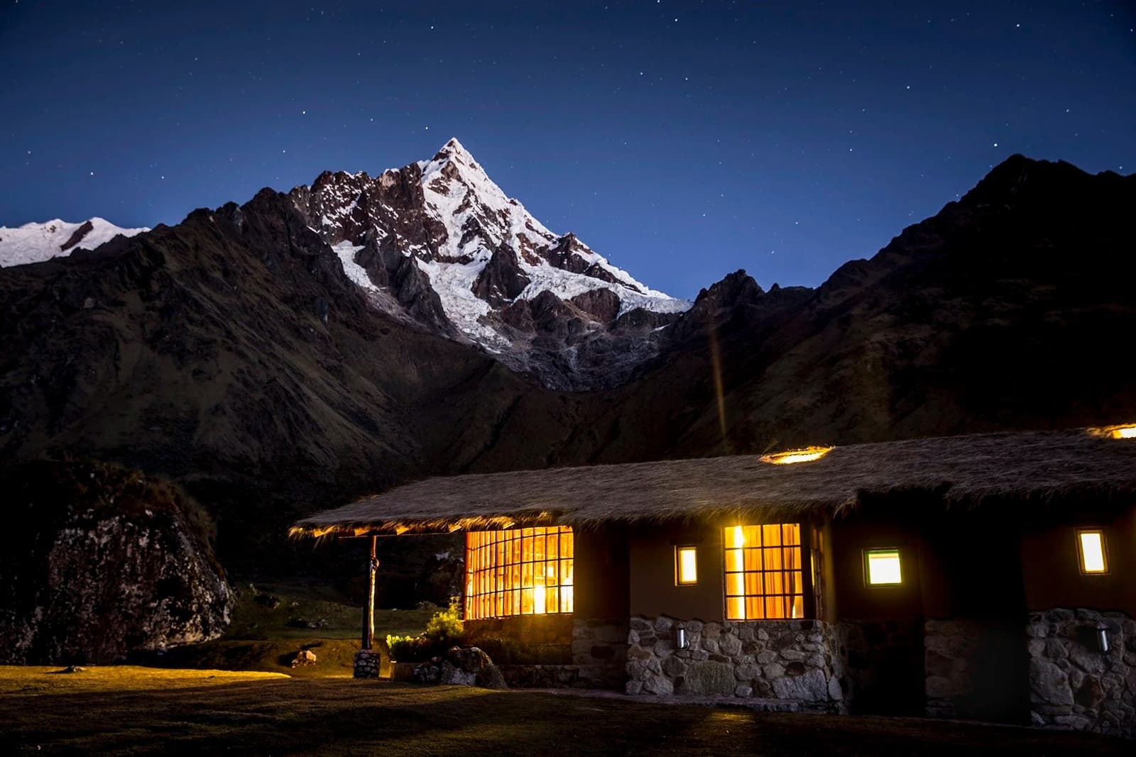 A hot tub waits inside, the glacier does not care, and somehow both feel right. A warmly lit stone-and-thatch mountain lodge at night during an andes trek, with a snow-capped Andean peak rising under a star-filled deep blue sky