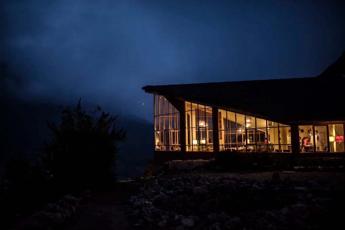 Cold air outside, warm light within, the lodge earns its keep after a long day on the trail. A mountain lodge glowing with warm interior lights against a deep blue dusk sky on an andes trek, with mist rolling in over the surrounding hills