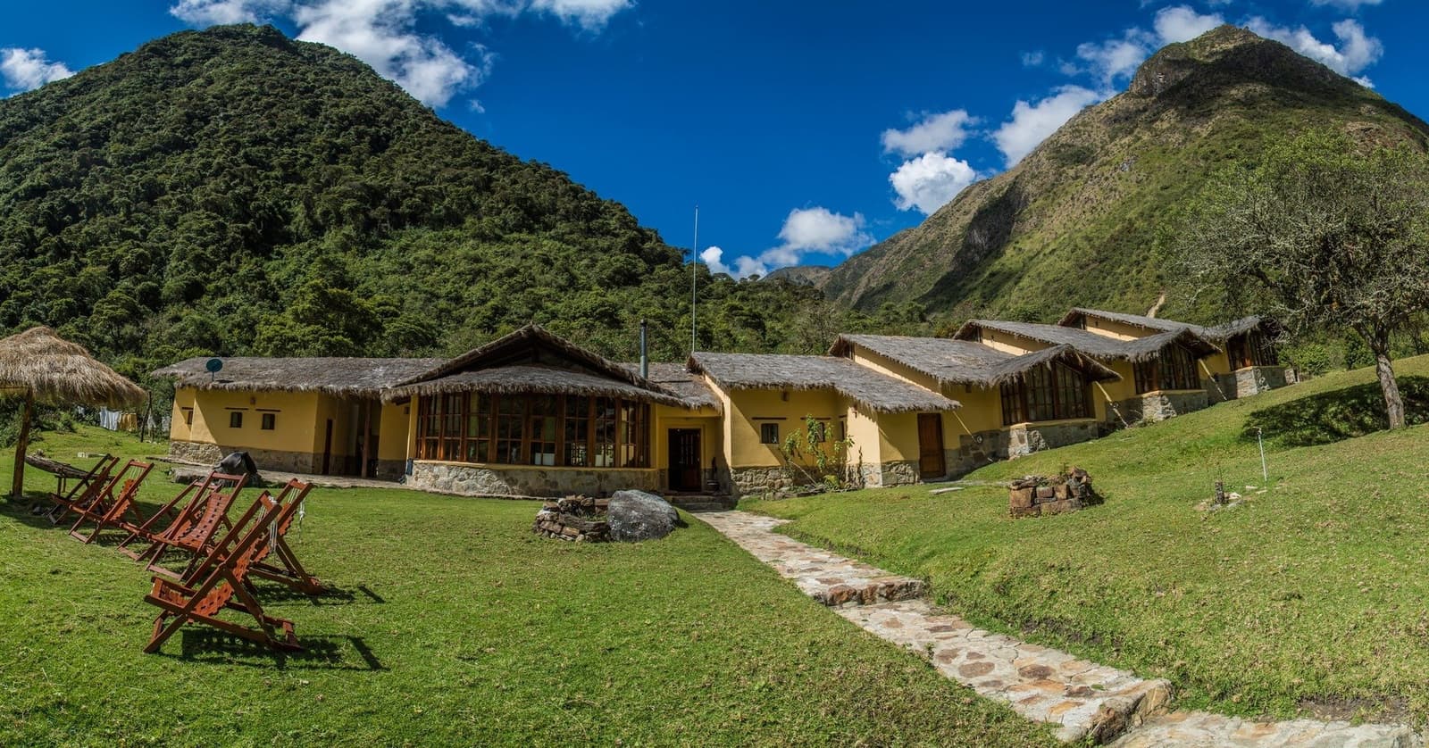 Your base camp for the night, and the mountains are included at no extra charge. Yellow-walled mountain lodge with thatched roofs and wooden deck chairs on a green lawn, set between two steep forested peaks during an Andes trek in Peru