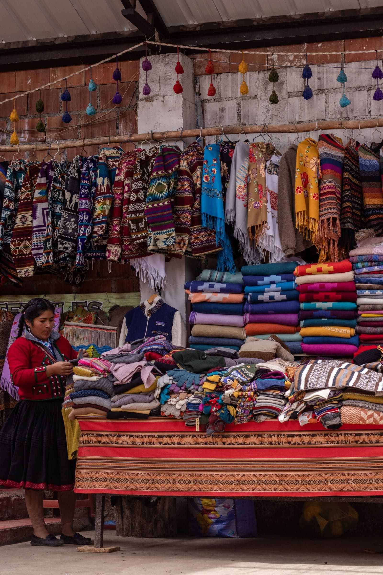 Every color the Andes has to offer, folded and hanging within arm's reach. Photo: leandra favan A woman in traditional Andean dress arranges folded wool textiles at an open-air market stall, with rows of patterned ponchos and embroidered scarves hanging overhead and stacks of colorful blankets piled high on the table — a common stop on an andes trek through the Sacred Valley