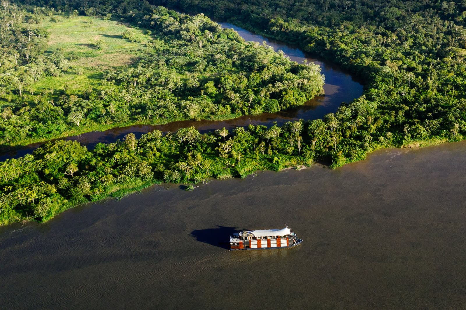 Small ship, very big forest. The scale only makes sense from up here. Photo: Tanveer Badal Aerial view of the Aria Amazon cruise ship navigating a wide dark-water river past dense rainforest and a narrow jungle tributary in Peru