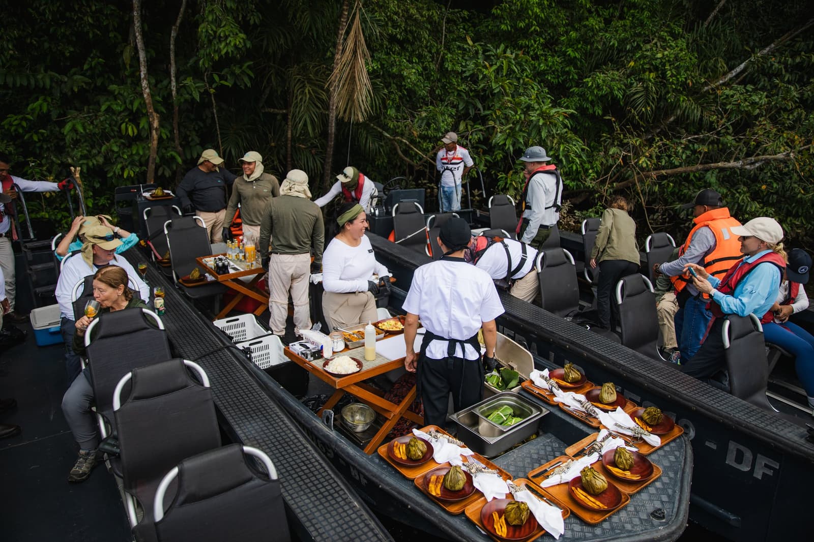 Lunch delivered dockside, deep in the forest, with a full kitchen crew and nowhere else you'd rather be. Photo: Yvonne Kreuzmayr Staff aboard the Aria Amazon cruise serve a spread of traditional Amazonian dishes on wooden trays between two skiffs moored at a dense rainforest riverbank, with guests seated and crew in white uniforms presenting food wrapped in banana leaves