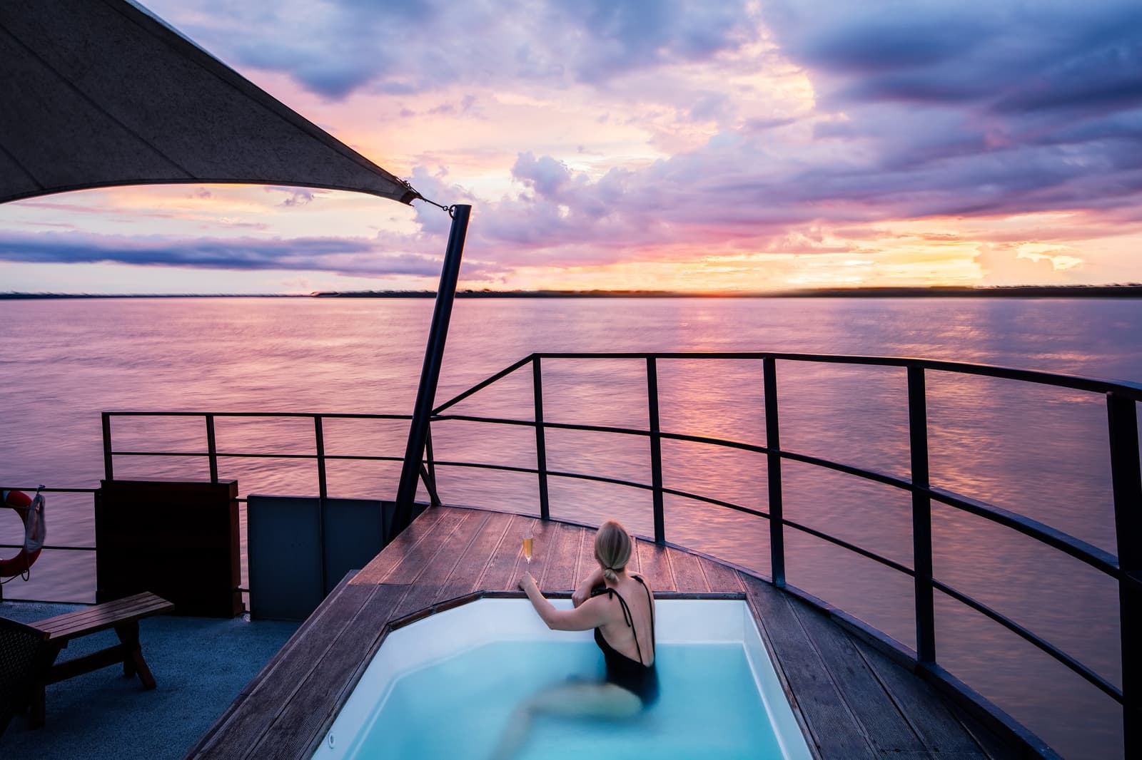 Sundowners hit different when the river is your horizon. Photo: Scott Sporleder A woman soaks in the outdoor jacuzzi on the upper deck of the Aria Amazon cruise ship, holding a glass of sparkling wine while a vivid pink and orange sunset reflects across a wide Amazonian river