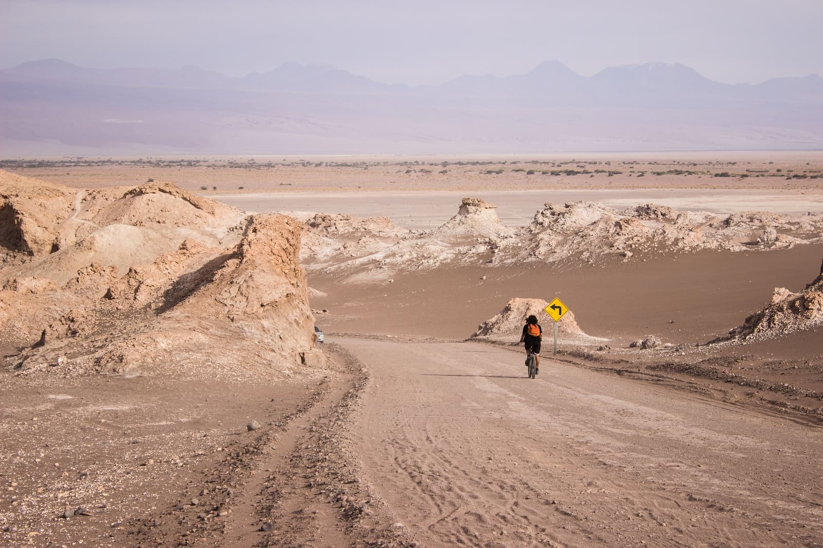 Tú, el polvo y una señal que confirma la curva. Photo: Bike Atacama Un ciclista con mochila naranja pedalea por un camino de tierra entre las formaciones de sal y roca erosionadas del Valle de la Luna durante una expedición de camping en el desierto de Atacama, con cumbres andinas difuminadas en la neblina al fondo.