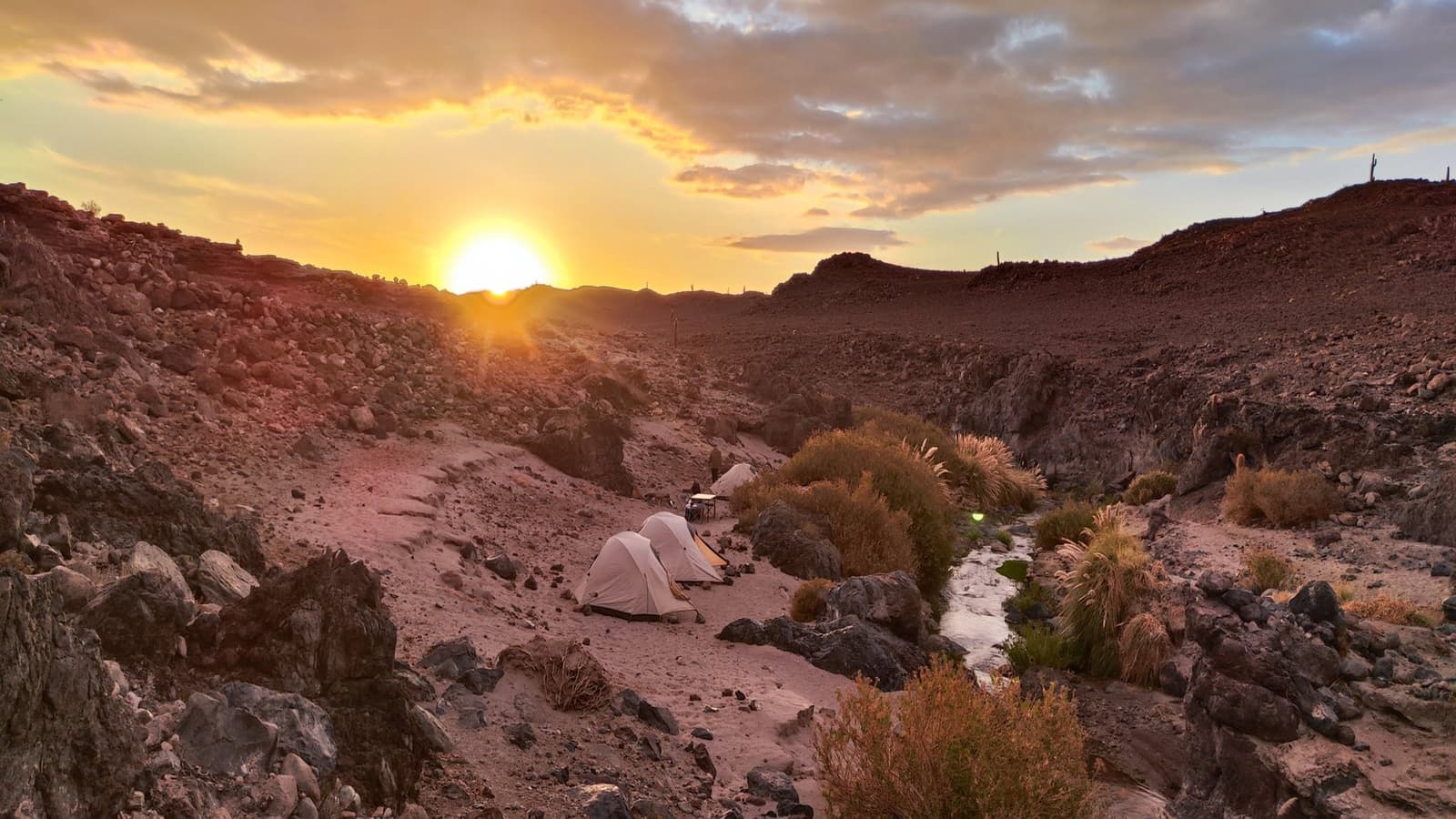 La quebrada te da techo, el sol te da despedida: eso es acampar en el Atacama. Tres carpas tipo domo instaladas en una quebrada rocosa durante un camping en el desierto de Atacama, con un pequeño arroyo y arbustos secos cercanos mientras el sol se pone entre dos crestas, bañando el terreno volcánico rojizo de luz dorada