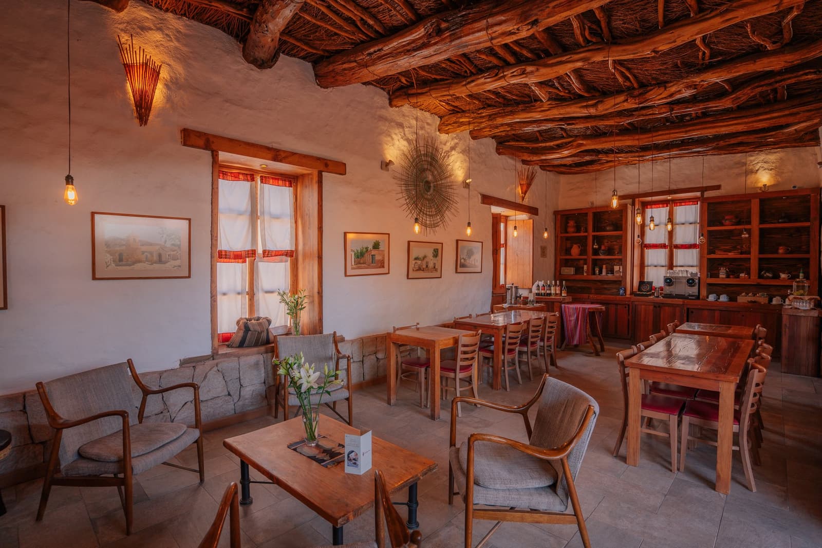 Flores frescas, luz cálida y silla de verdad: el desierto puede esperar un rato. Acogedora sala comedor de adobe con techo rústico de troncos, lámparas colgantes Edison, mesas y sillas de madera, acuarelas locales y una decoración de sol tallado en la pared de la lodge base para una expedición de camping en el desierto de Atacama