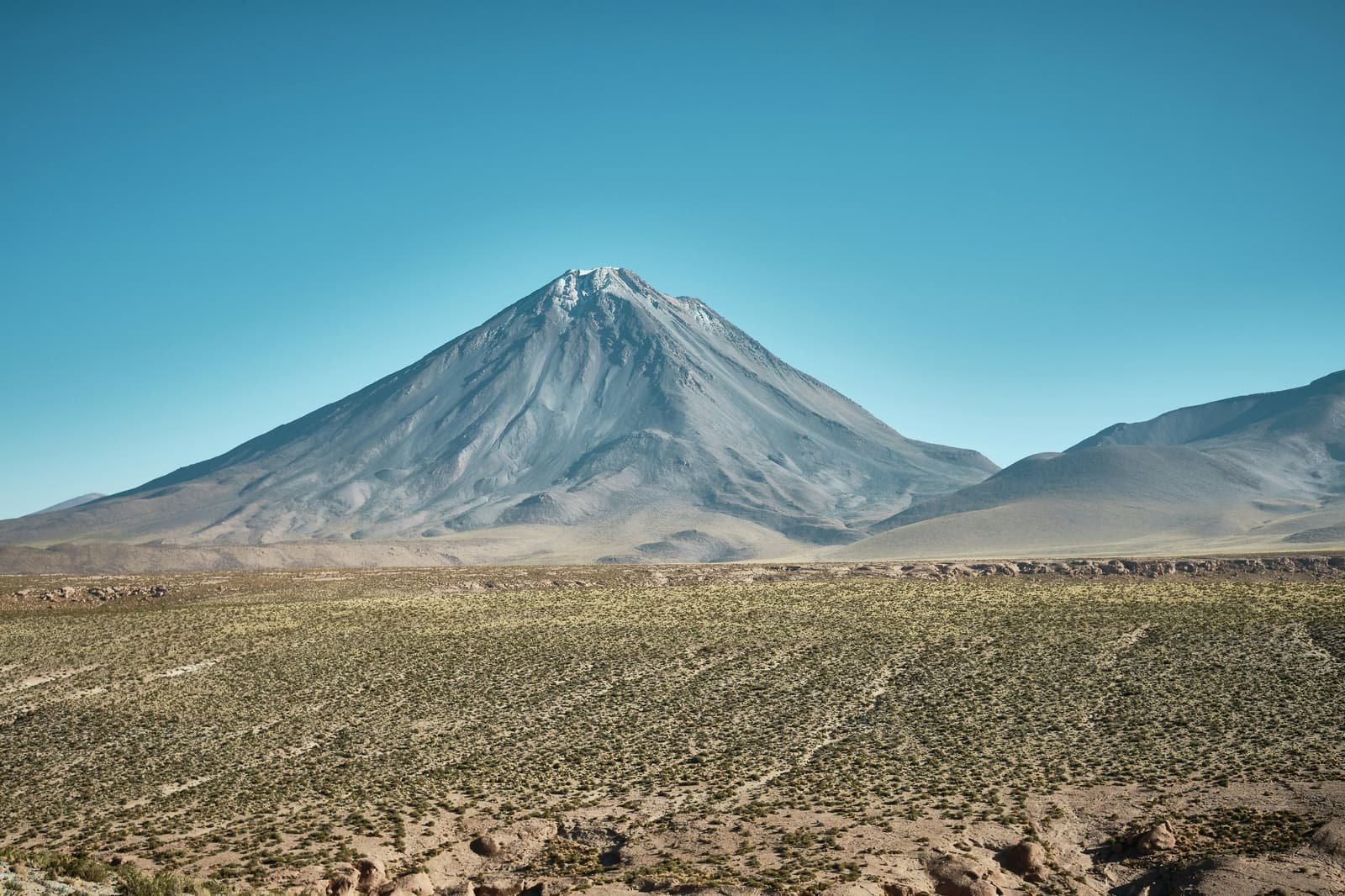 Un cono tan limpio como este no sale en una tarde. Un volcán de forma cónica perfecta con cima nevada se eleva sobre una llanura altiplánica de vegetación escasa bajo un cielo azul despejado, en el paisaje de camping en el desierto de Atacama.