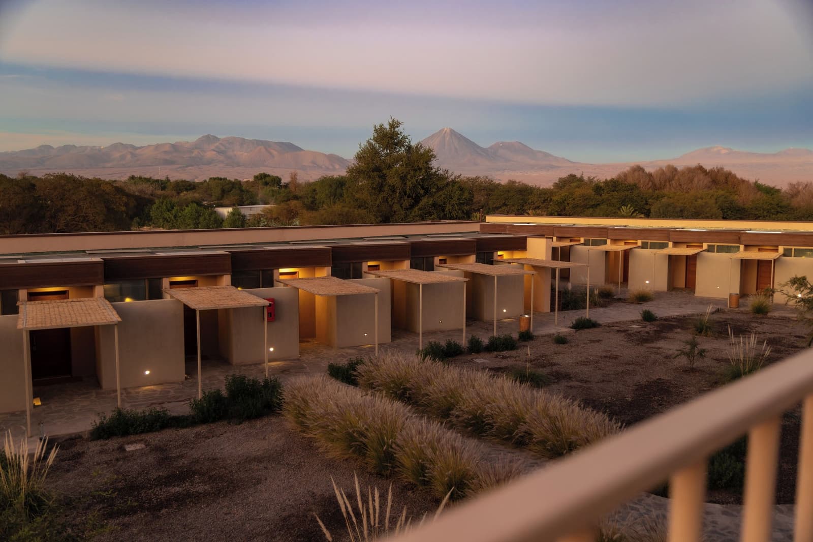 Your room for the next three nights, with a volcano keeping watch. Photo: Cinco_Estudio Low-slung adobe bungalows with warm pathway lighting in an atacama desert tour package hotel courtyard, native grasses in the foreground and a cone-shaped volcano rising above the treeline at dusk