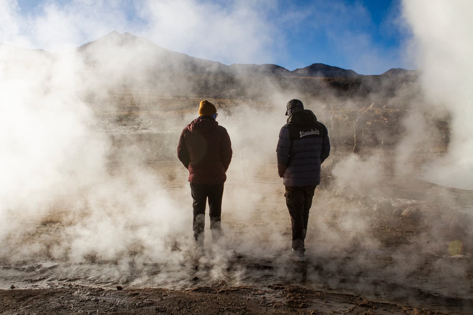 Cold face, warm ground, and the earth reminding you who's in charge. Photo: Tatio Caminata Two travelers in heavy winter jackets stand amid thick geothermal steam plumes at El Tatio geysers, part of this atacama desert tour package, with volcanic peaks visible under a blue sky in the background