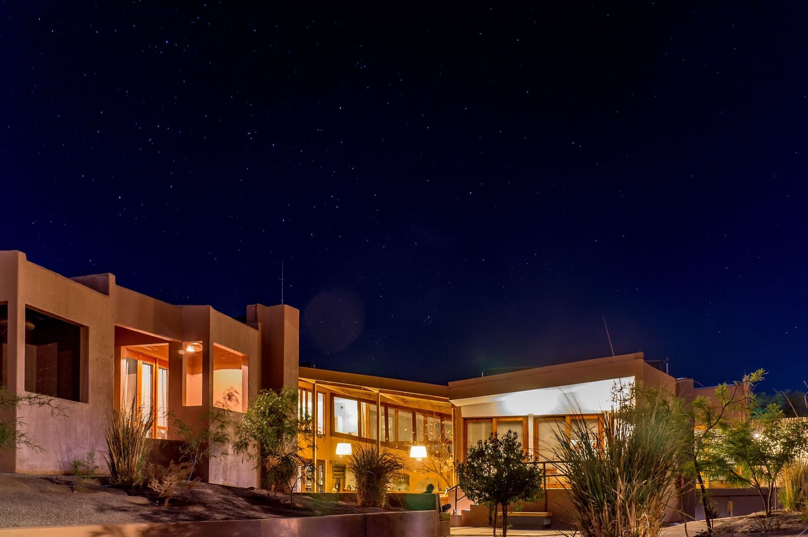 Stars out, lights on, and the desert is already doing its thing. Exterior night view of a low-rise adobe-style lodge illuminated by warm interior lights, with a clear star-filled sky overhead, part of an Atacama desert tour package stay in San Pedro de Atacama