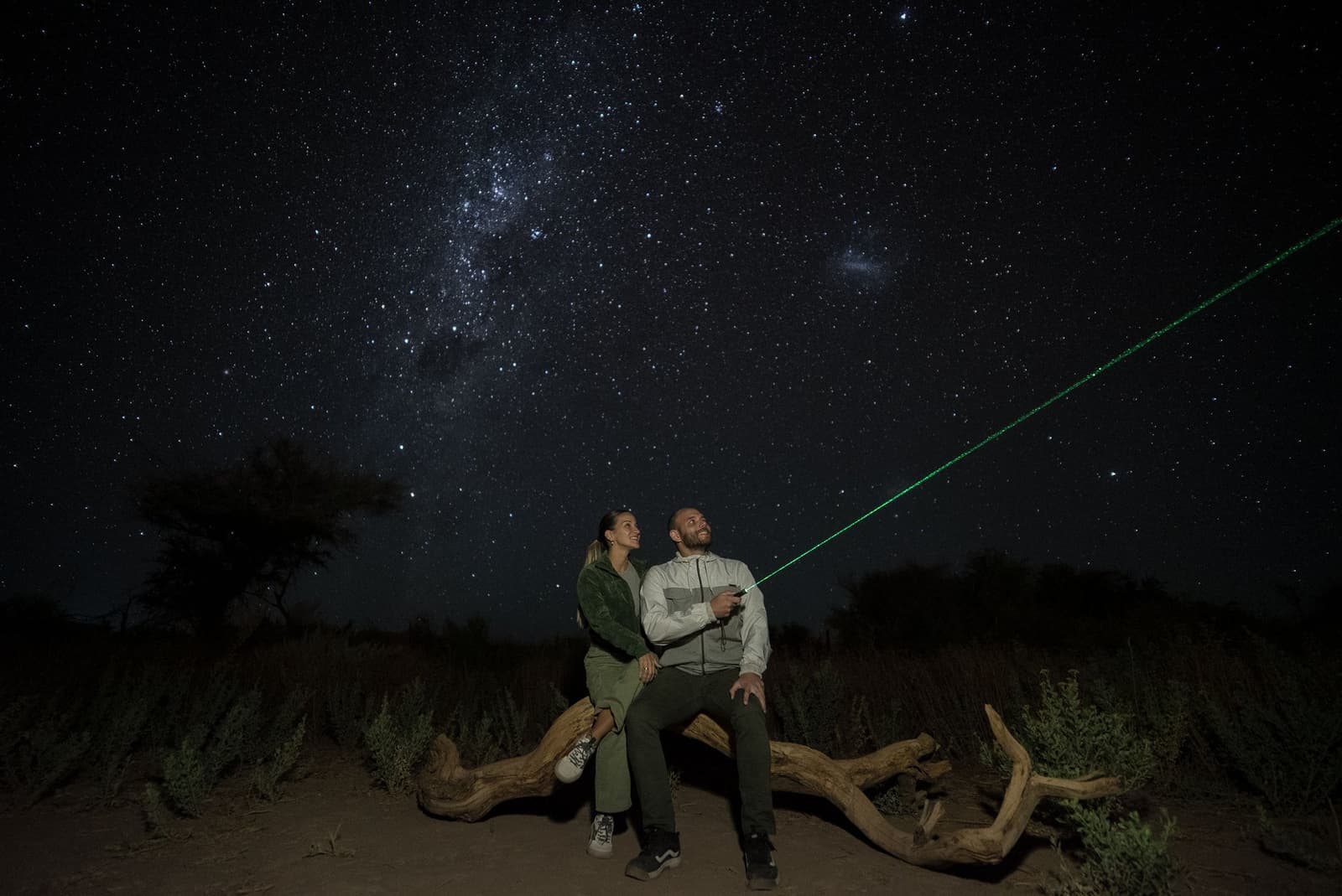 One green laser, two people, and more stars than you can count. A couple sitting on a driftwood log in the Atacama Desert, smiling as a green laser pointer traces a path across the Milky Way on a clear stargazing night