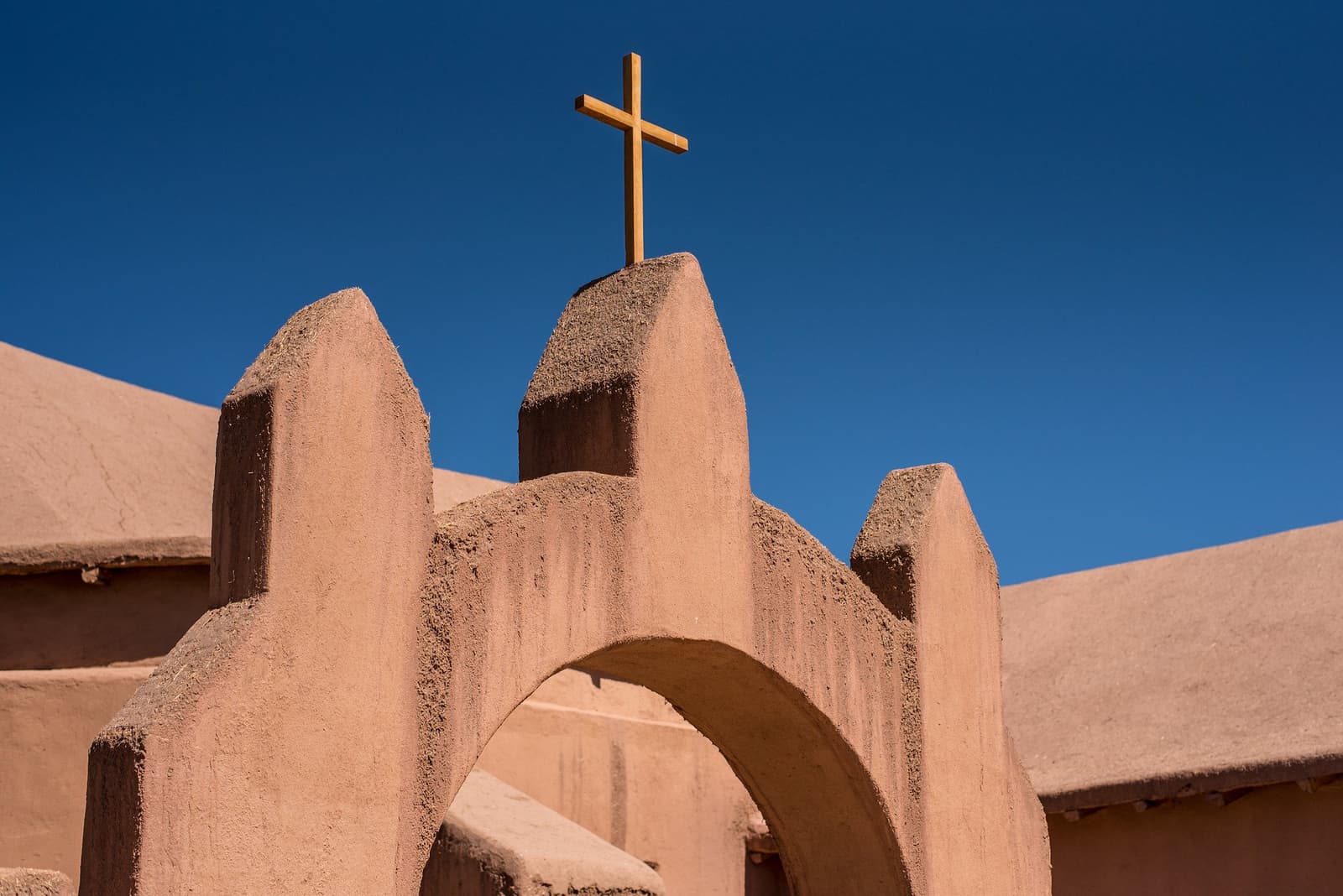 Eeuwen woestijnzon, en dit kerkje staat er nog net zo kalm bij. Close-up van een rustieke adobe kerkgevel met kantelen en een houten kruis tegen een diepblauwe lucht