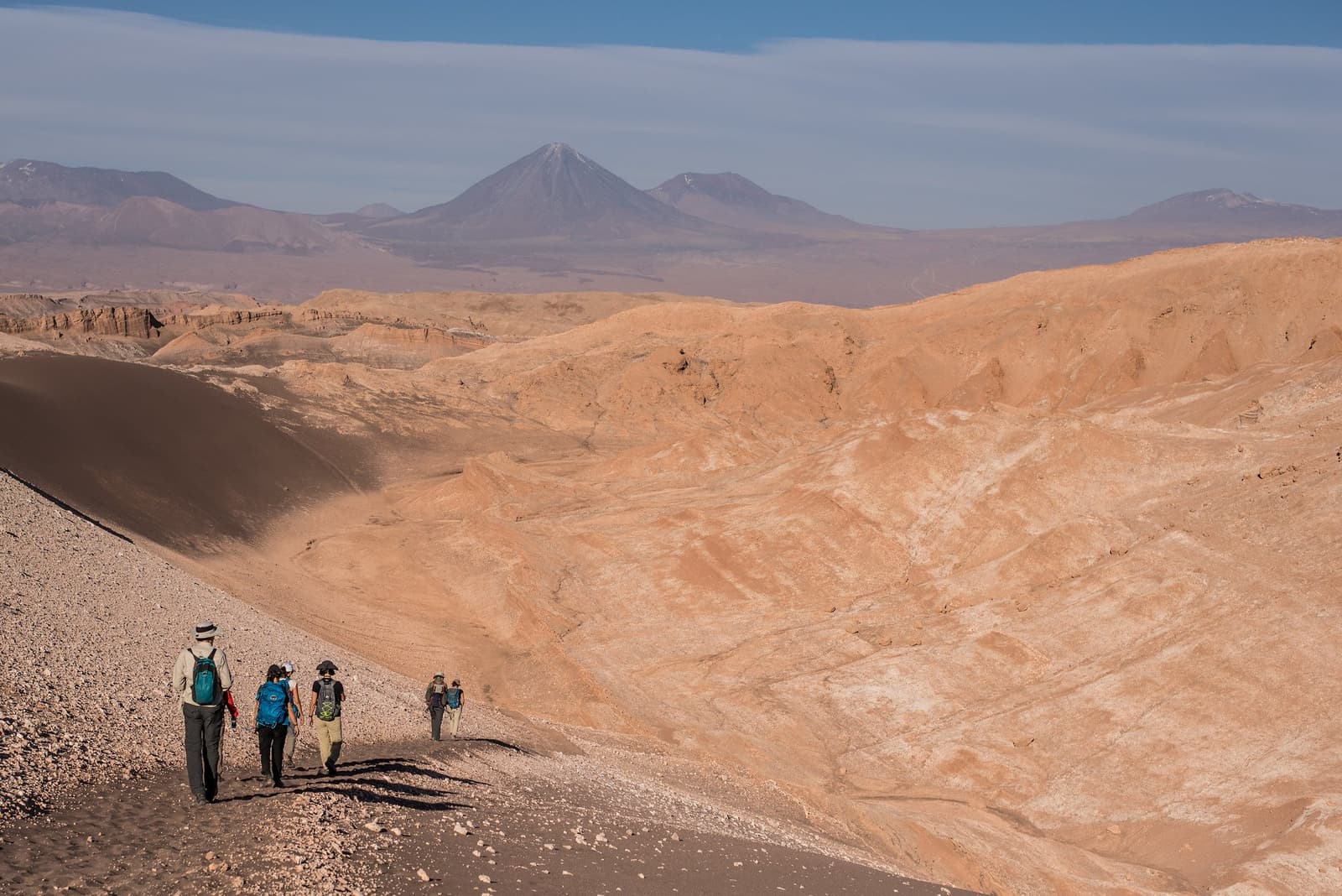 Vijf mensen, één immens landschap, en nergens anders te zijn. Photo: Moon Valley Hike Een groep van vijf wandelaars met rugzakken daalt een rotsachtige rug af tijdens een Atacama woestijntrek door de Maanvallei, met uitgestrekte okerkleurige badlands en twee besneeuwde vulkaantoppen aan de horizon.