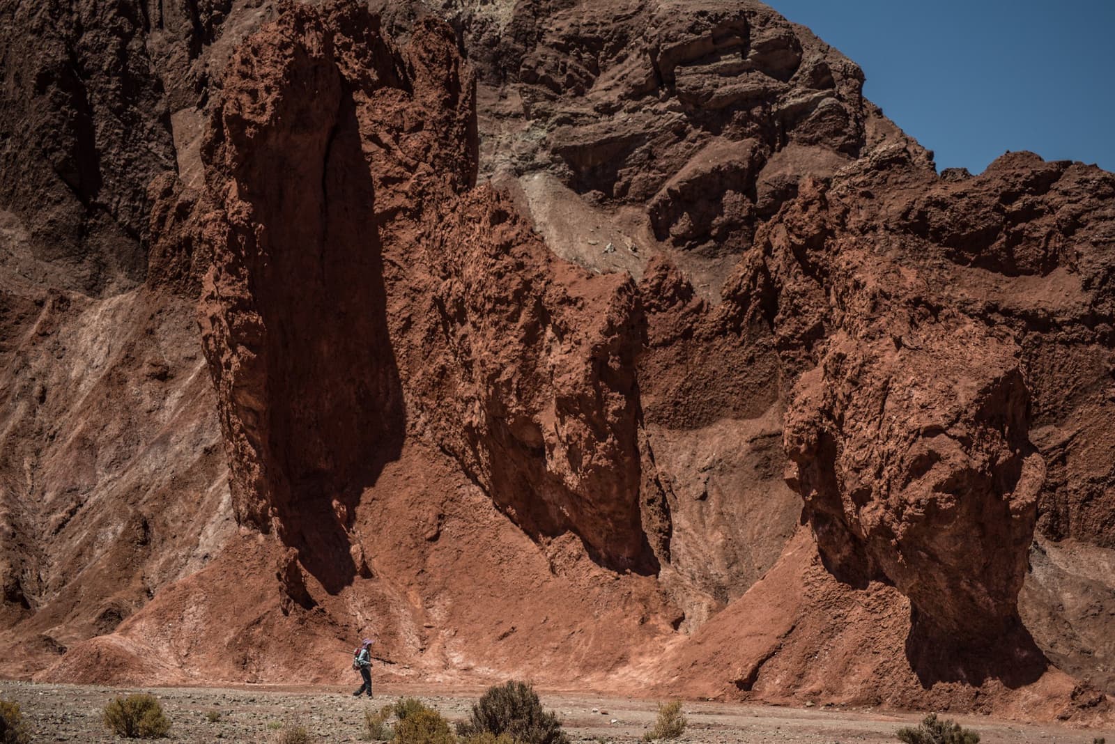 Zo klein, zo'n mens. Zo groot, zulke rotsen. Photo: Rainbow Valley Een eenzame wandelaar loopt onder hoge rode en koperkleurige geërodeerde rotsformaties tijdens een Atacama Desert Trek, met schaarse woestijnstruiken op de voorgrond en een strakblauwe lucht.