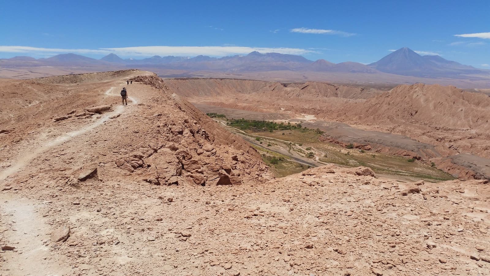 Eén richel, een heel gebergte, en zoveel stilte als je maar wilt. Een wandelaar met rugzak loopt over een smalle rotsachtige richel tijdens een Atacama woestijn trek, met een groene vallei diep beneden en een rij besneeuwde Andesbergen aan de horizon