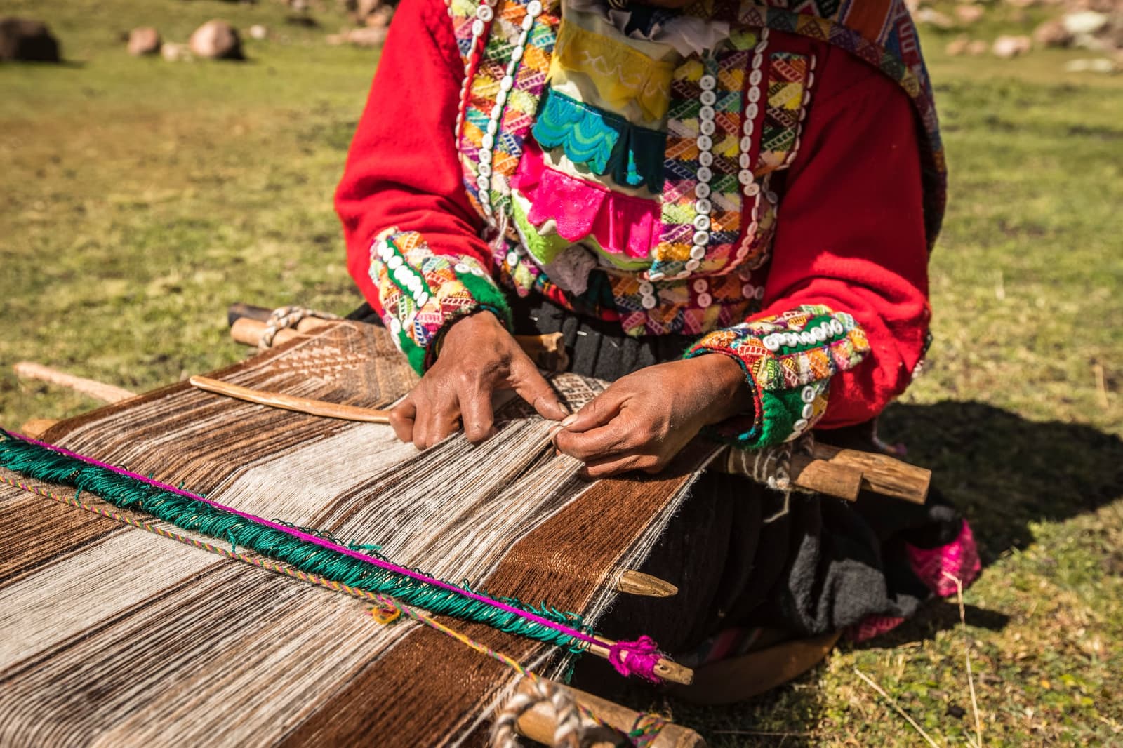 Centuries of pattern and memory, passed down through hands like these. Photo: CH DECLERCQ A woman in traditional colorful Andean dress works a backstrap loom on open grassland during the Ausangate Lodge Trek, her hands guiding brown and cream alpaca wool threads with a wooden shuttle