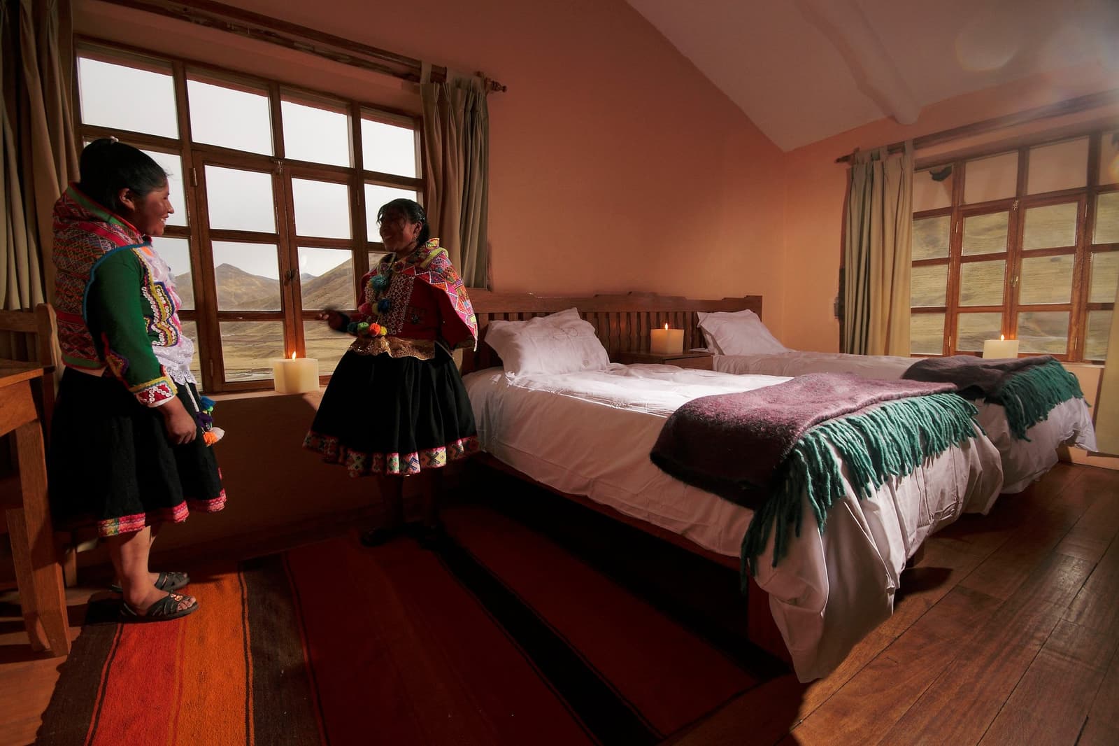 Your hosts know exactly which blanket you'll reach for first. Photo: Gustavo Vivanco Leon Two Andean women in traditional woven dress show guests into a candlelit twin-bed lodge room on the Ausangate Lodge Trek, with high-altitude peaks visible through wooden-framed windows