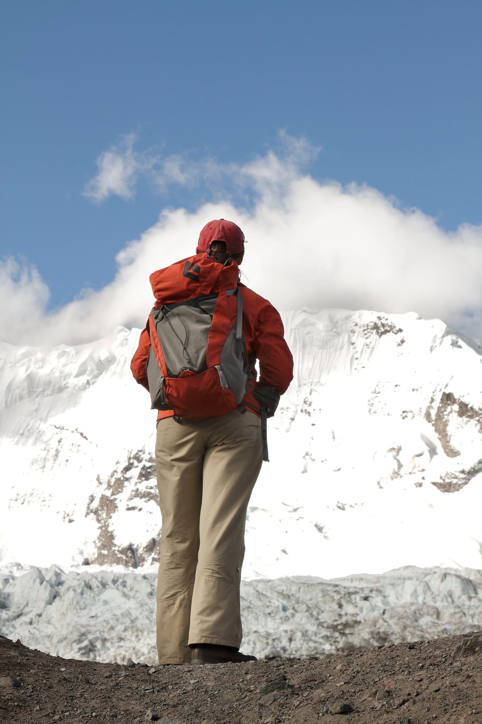 Up here, the mountain does the talking. You just listen. Photo: Gustavo Vivanco Leon A hiker in a red jacket and backpack stands on rocky moraine facing a massive snow-covered peak on the Ausangate Lodge Trek, with clouds wrapping the summit and glacier ice visible at its base