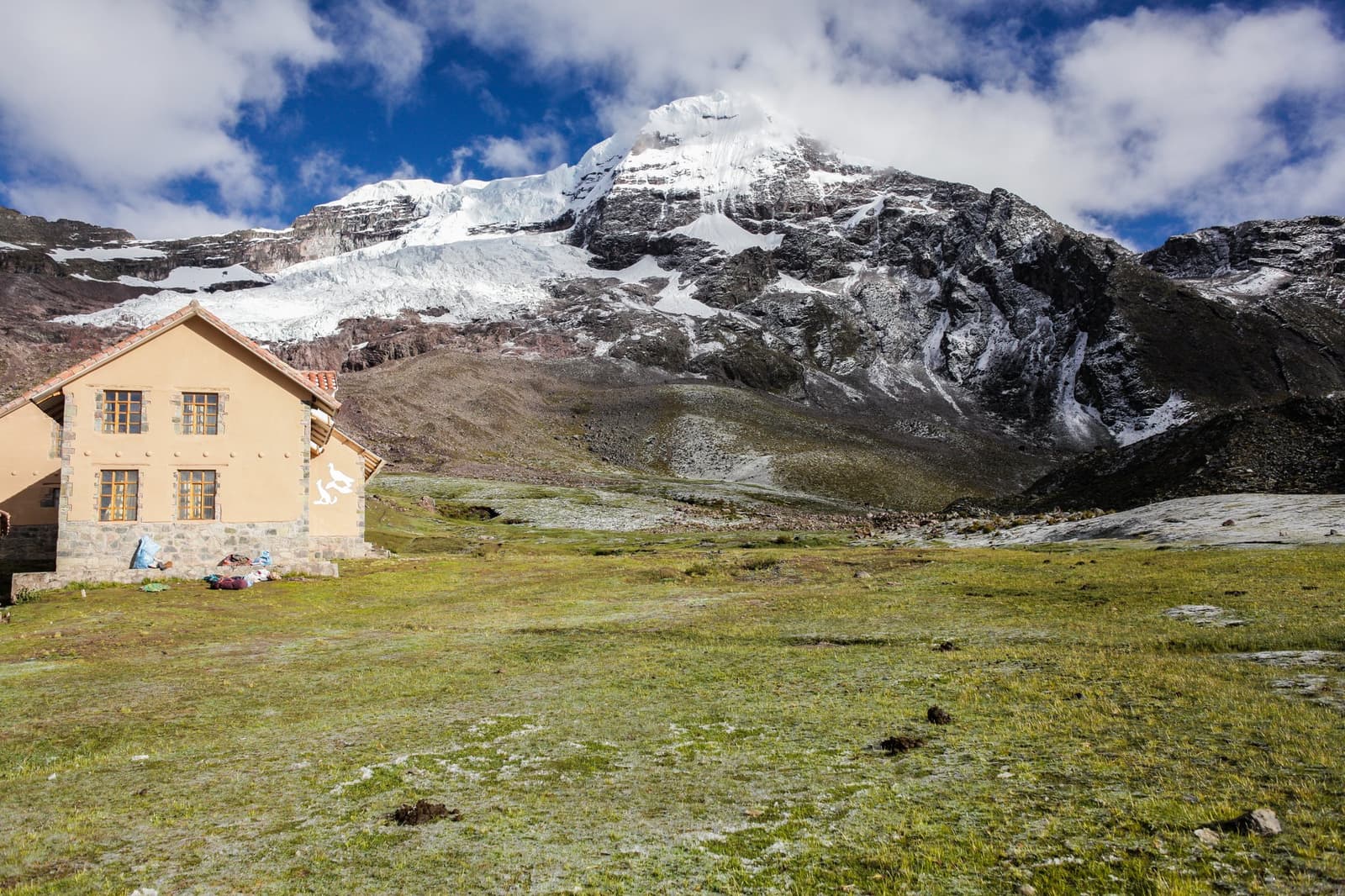 Tonight's base camp has better beds than it looks, and that glacier isn't going anywhere. A two-storey stone and plaster mountain lodge sits on an open high-altitude grassland on the Ausangate Lodge Trek, with a heavily glaciated peak rising steeply behind it under a blue sky with scattered clouds