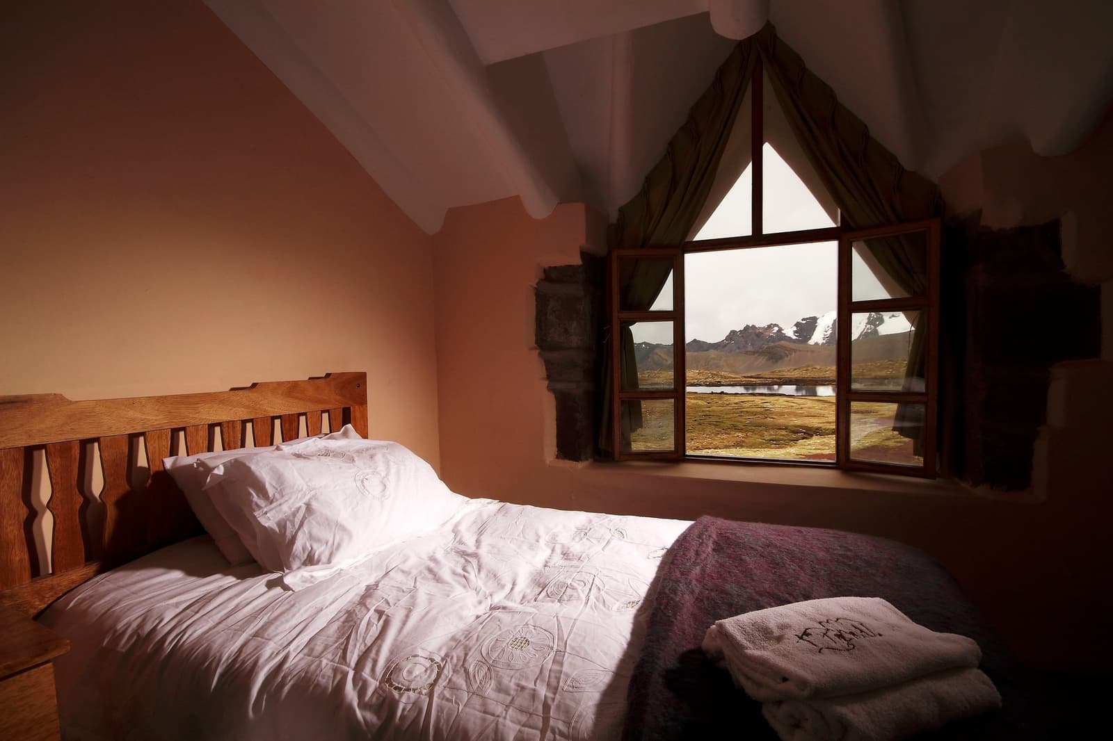 Sleep well at altitude. The mountains are right there when you open your eyes. Photo: Gustavo Vivanco Leon Cozy lodge bedroom on the Ausangate Lodge Trek, with a carved wooden headboard, white embroidered duvet, folded towels, and a triangular gabled window framing a high-altitude wetland and snow-dusted peaks beyond
