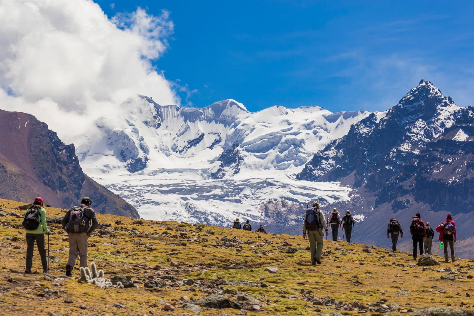 Dünne Luft, riesige Berge und eine Gruppe Menschen, die sich gut vorbereitet hat. Photo: Paul Spierenburg Eine Gruppe von Wanderern mit Rucksäcken und Wanderstöcken überquert eine felsige Hochlandwiese beim Ausangate Lodge Trek, im Hintergrund ein gewaltiger schneebedeckter Gletscher und gezackte Andengipfel unter blauem Himmel