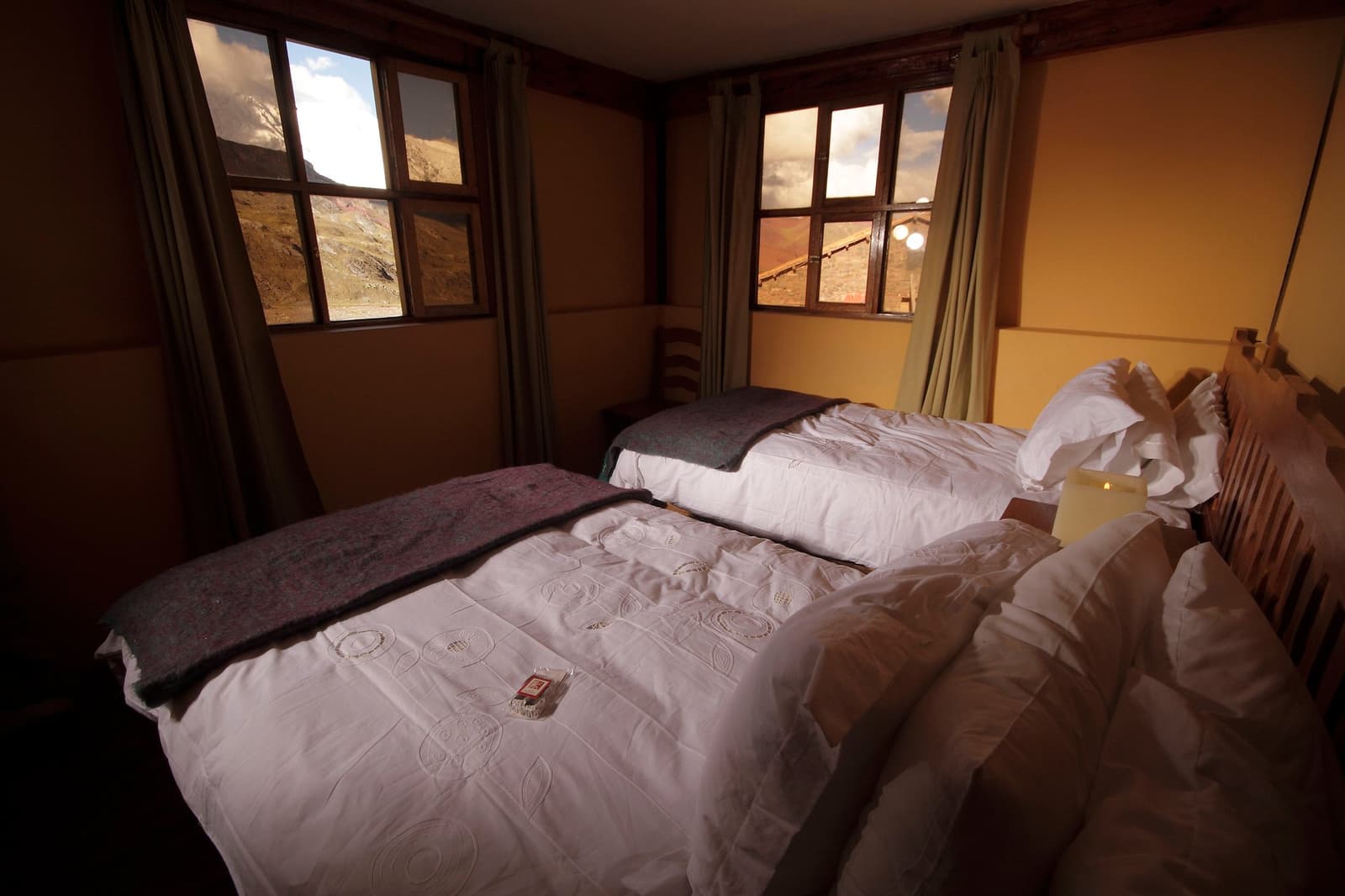 Wool blankets, crisp linen, and a mountain outside your window. Rest earned. Photo: Gustavo Vivanco Leon A twin-bedded mountain lodge room on the Ausangate Lodge Trek, with white embroidered duvets, wool blankets folded at the foot of each bed, and two windows framing rugged Andean peaks outside