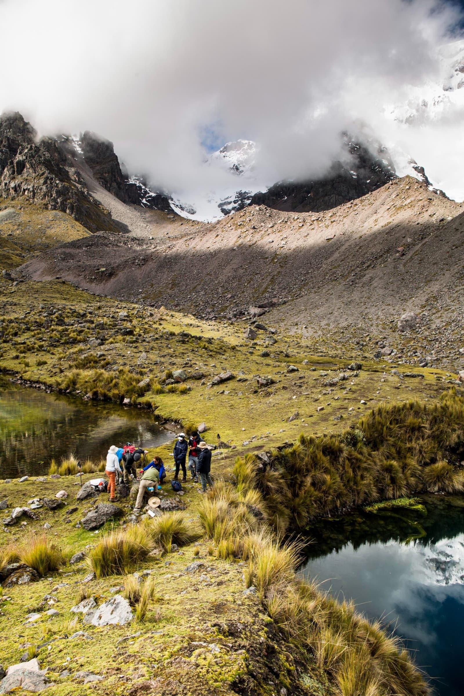 A good excuse to stop, catch your breath, and let the mountain do the talking. A group of trekkers rests beside a high-altitude alpine lake on the ausangate trek, with ichu grass, mossy boulders, and a glaciated peak wrapped in clouds rising steeply behind them