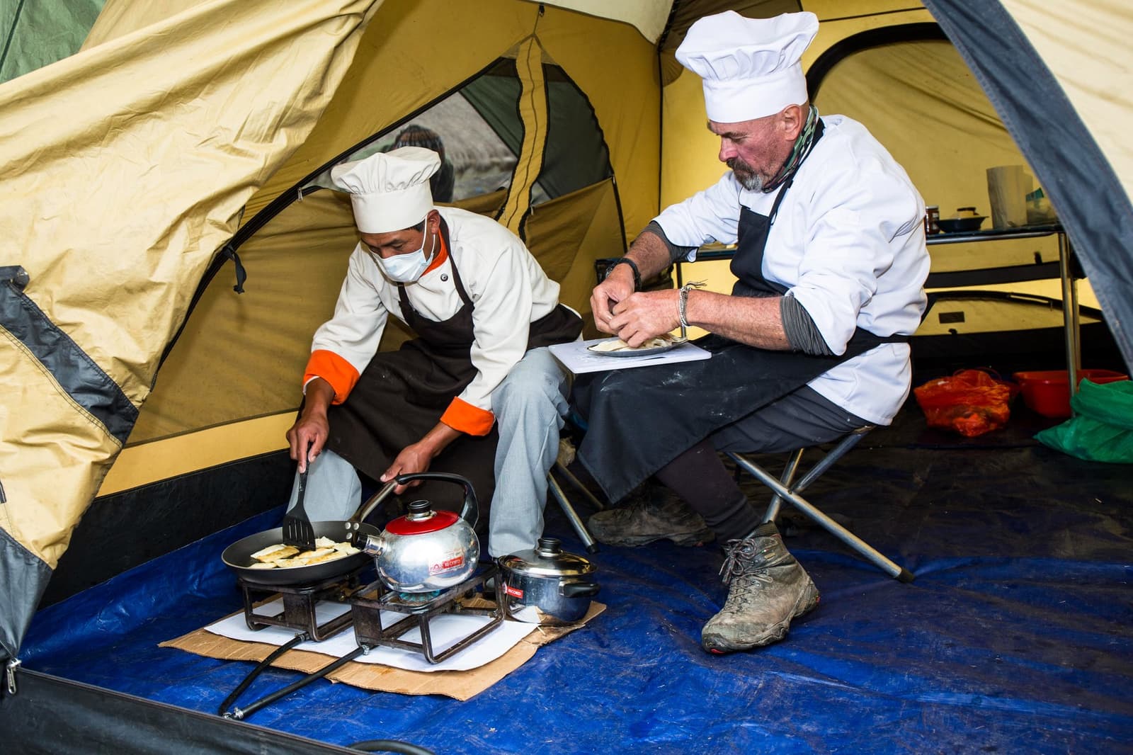 Full chef whites, a camp stove, and no complaints about the kitchen size. Two cooks in white chef hats and aprons prepare food on a portable gas stove inside a yellow camping tent on the Ausangate trek, with a kettle, frying pan, and plated dish in hand