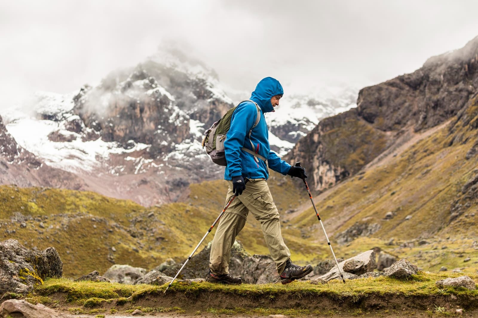 Poles out, hood up, one foot in front of the other. Some days the mountain asks that of you. A hiker in a blue hooded jacket and backpack uses trekking poles along a rocky high-altitude trail on the Ausangate trek, with snow-dusted peaks and mist-covered cliffs filling the background
