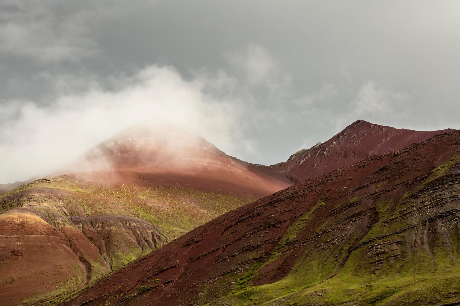 The mountain picks its own mood, and today it chose crimson. Deep red and burgundy mineral peaks on the Ausangate trek, with low clouds wrapping one summit and patches of bright green ichu grass on the steep slopes