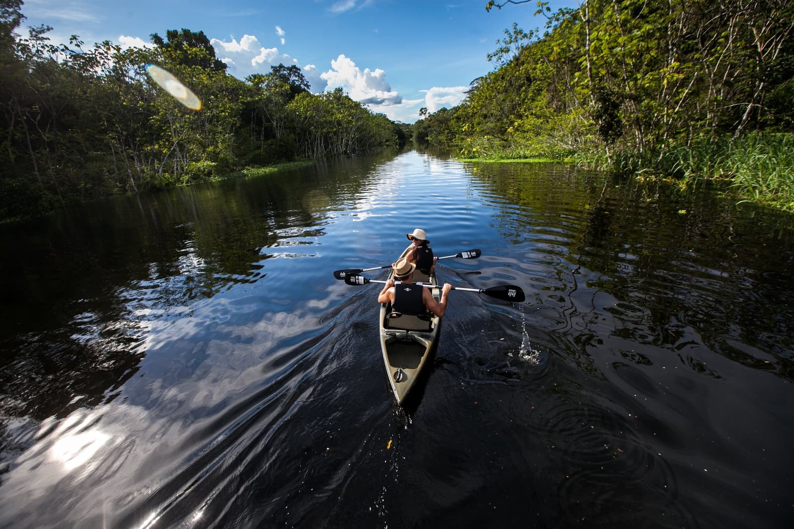 Água quieta, quatro braços e um rio que recompensa quem não tem pressa. Photo: er: Rodrigo Rodrich Dois viajantes remam um caiaque duplo por um canal de água preta calmo, ladeado por densa floresta amazônica, em uma excursão do Delfin Amazon Cruise