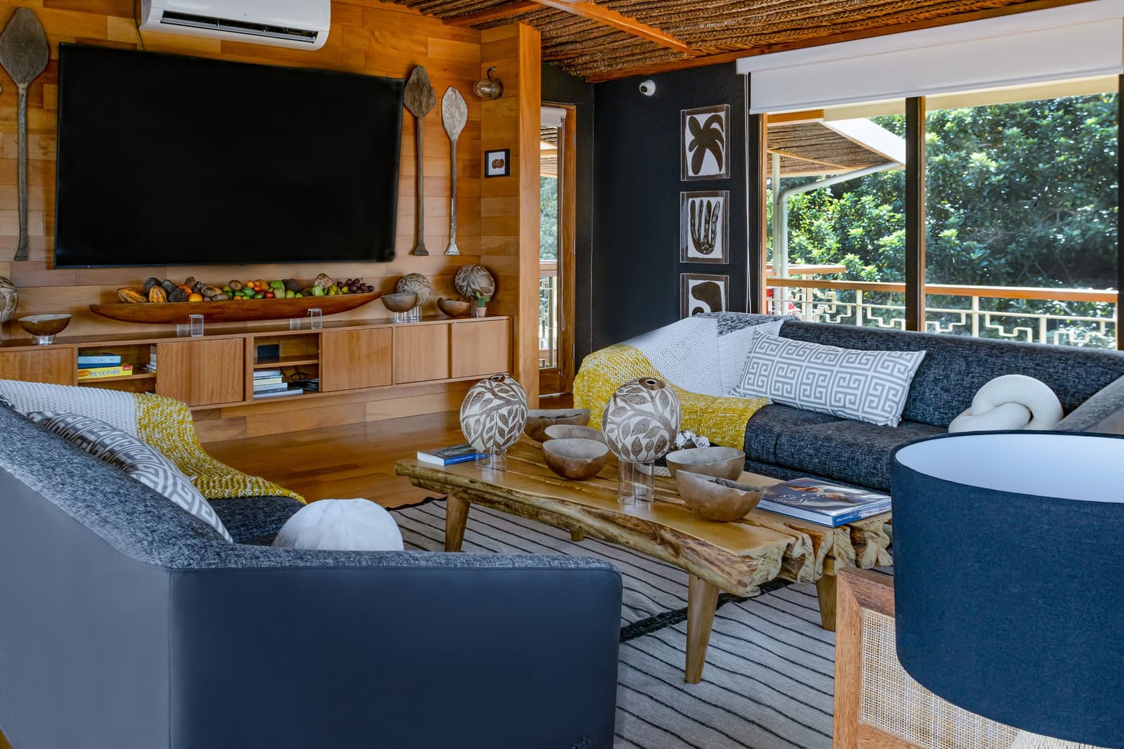 Livros na mesa, a floresta logo ali do outro lado do vidro. Difícil querer ir embora. Área de lounge confortável a bordo do cruzeiro Delfin na Amazônia, com sofás azul-escuro, mesa de centro em madeira bruta decorada com esferas de cerâmica artesanal e tigelas de madeira, paredes de madeira aquecidas, obras de arte botânicas e uma grande janela com vista para a floresta tropical