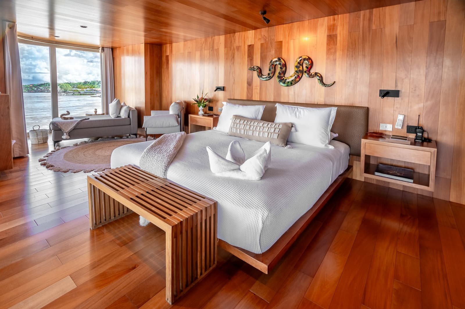 Madeira, linho branco e o rio passando pela janela. O Amazonas nunca coube tão bem num quarto. Photo: Mario Gomi Suíte master espaçosa no cruzeiro Delfin Amazon com painéis de madeira aquecida, cama king com roupa branca, escultura colorida de cobra na parede e janela do chão ao teto com vista para o rio