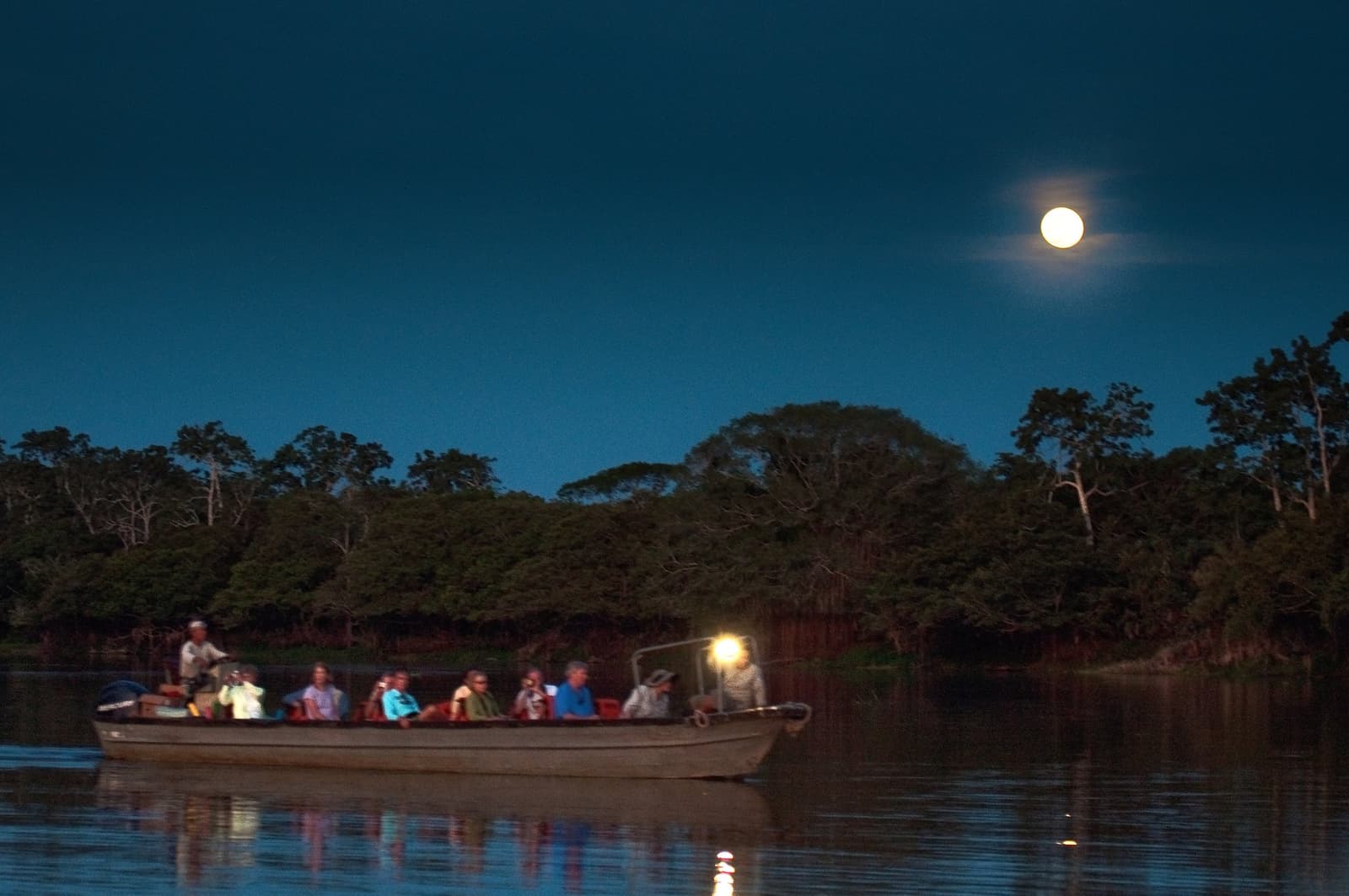 O rio à noite tem sua própria lógica, e a lua mostra o caminho. Um grupo de viajantes desliza em um barco de madeira por um rio escuro durante um cruzeiro Delfin pela Amazônia, com uma lua cheia brilhante surgindo acima da copa das árvores sob um céu crepuscular azul profundo
