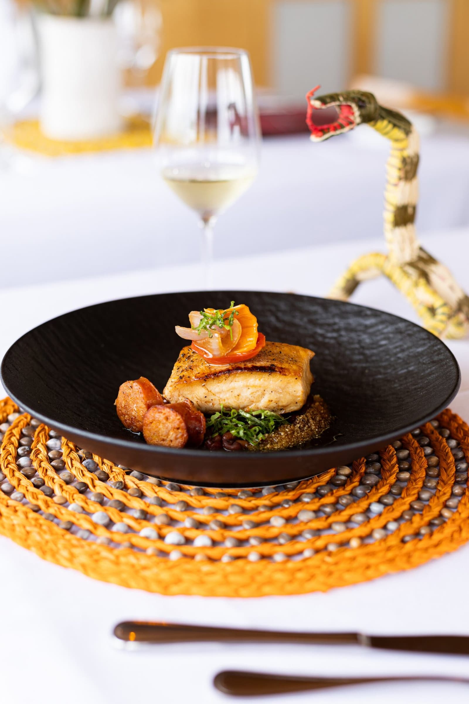 Ingredientes regionais, técnica de verdade, e uma cobra decorativa de guarda sobre a mesa. Filé de peixe selado em tigela escura com legumes salteados e chouriço, servido sobre jogo americano de palha laranja ao lado de uma taça de vinho branco a bordo do cruzeiro Delfin pelo Amazonas