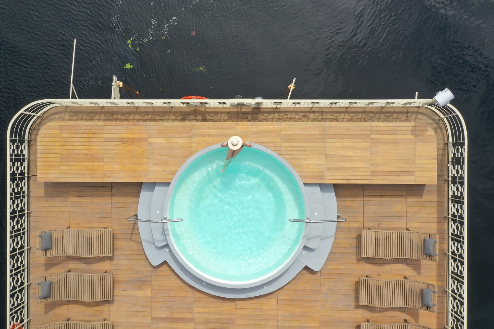Piscina lá em cima, rio em todo lugar, uma troca que vale a pena. Vista aérea de cima do convés de sol do Delfin Amazon Cruise, com uma piscina circular turquesa embutida em deck de teca, uma hóspede de chapéu branco na borda e as águas escuras da Amazônia visíveis além da grade