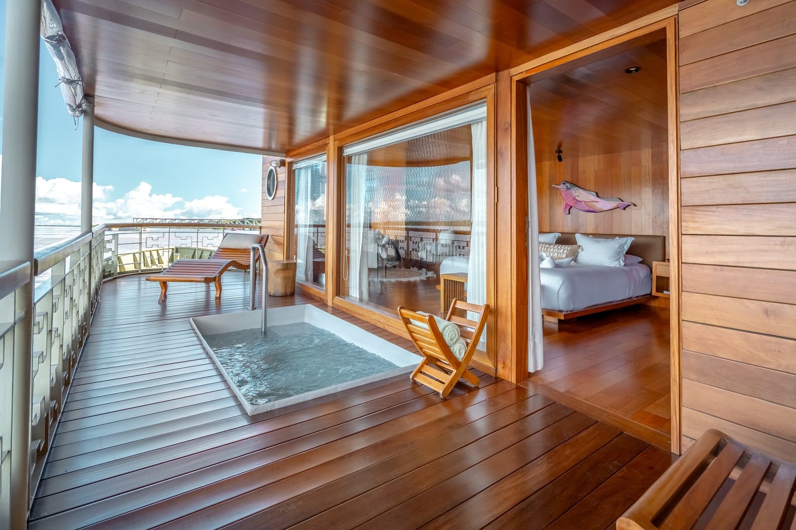 Jacuzzi próprio, o rio passando lá fora, o resto pode esperar. Photo: Mario Gomi Deck privativo de teca de uma suíte do cruzeiro Delfin Amazon com jacuzzi embutido, espreguiçadeira de madeira e toalhas dobradas sobre cadeira dobrável, com portas de vidro que abrem para um quarto com obra de arte de boto-cor-de-rosa na parede