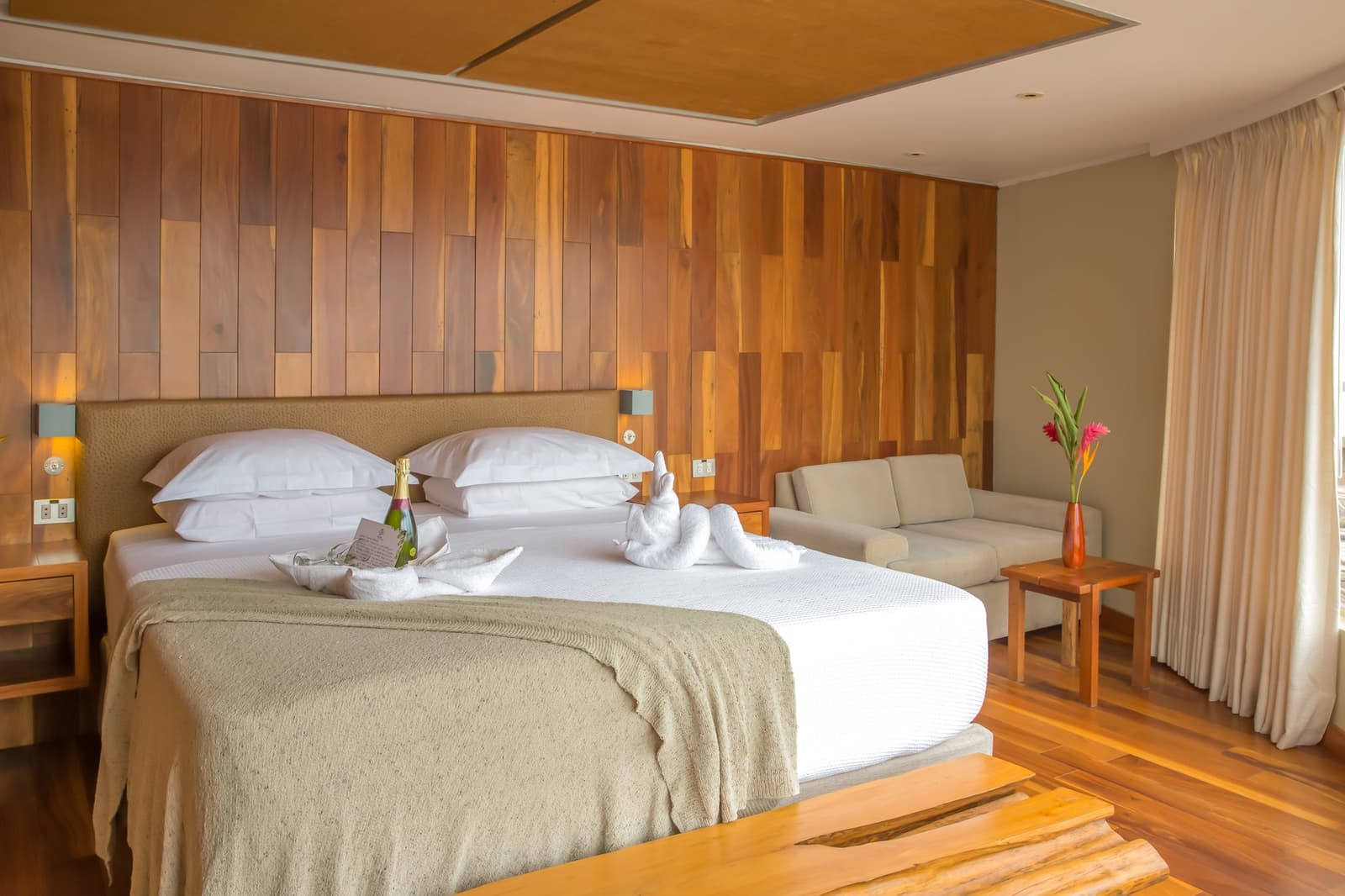 Um quarto que prova que luxo e floresta amazônica combinam muito bem. Photo: DIEGO PEREZ Suite espaçosa no cruzeiro Delfin Amazon com cama king ornamentada com toalhas dobradas em formato de cisnes, uma garrafa de champanhe, parede revestida em madeira aquecida, piso em madeira, sofá e arranjo de flores tropicais