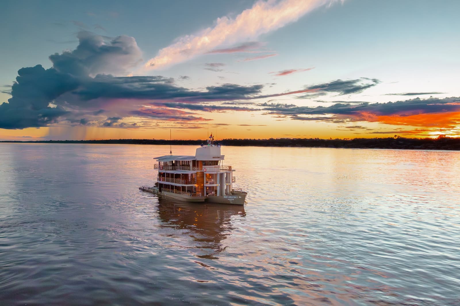 O drinque do fim do dia tem outro sabor quando o rio inteiro vira ouro. O navio de cruzeiro Delfin Amazon ancorado em um rio largo ao pôr do sol, com nuvens laranja e rosa refletidas na água calma e uma silhueta escura de floresta no horizonte