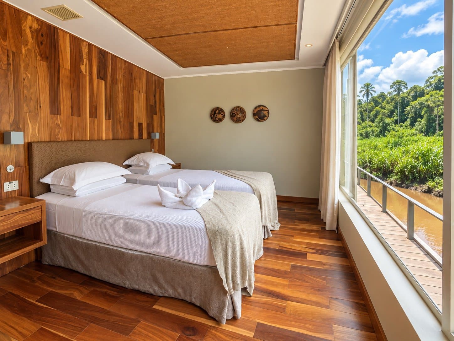 Roupa de cama branca, madeira quente e a Amazônia bem ali do lado. Suíte com duas camas no cruzeiro Delfin Amazon com piso de madeira nobre, parede revestida em madeira e janelas do chão ao teto com vista para a margem exuberante da floresta amazônica
