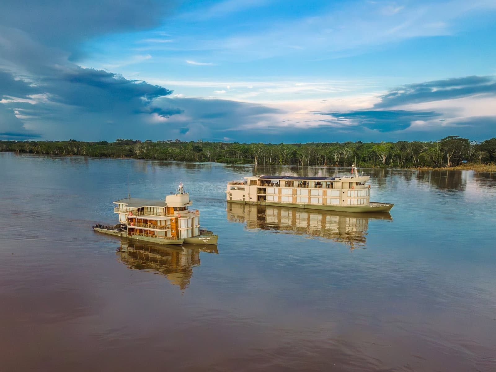 Dois navios, um rio, e uma reserva inteira esperando além das árvores. Vista aérea de dois navios do cruzeiro Delfin Amazon ancorados lado a lado em um amplo rio amazônico, com floresta inundada densa na margem oposta e nuvens de tempestade dramáticas em um céu azul