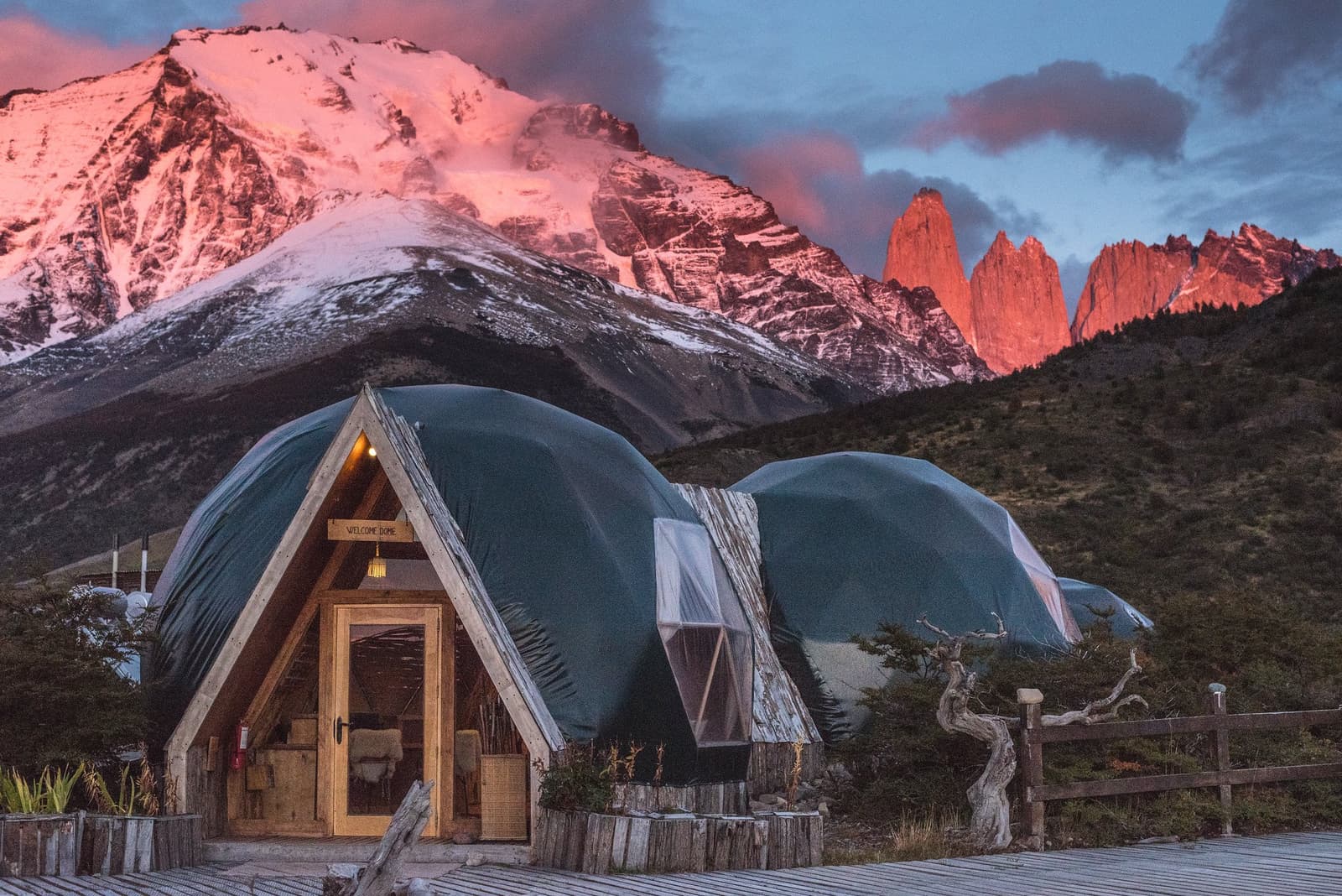 The welcome dome earns its name. Those towers behind it are your finish line. Illuminated geodesic welcome dome at EcoCamp Patagonia with the granite towers of Torres del Paine glowing deep orange at sunset behind snow-dusted peaks, part of the O trek in Patagonia
