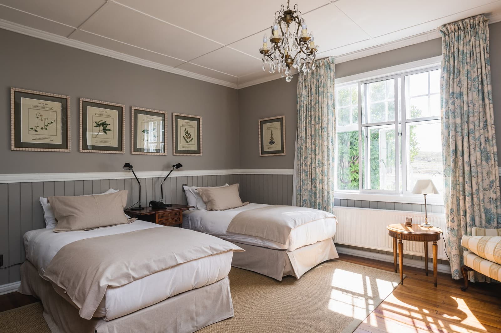Sunlight on the floorboards, botanical prints on the wall, the estancia does comfort without trying too hard. Photo: CARLOS DIAZ Twin room at Estancia Cerro Guido with two white-linen beds, a crystal chandelier, botanical prints on grey wainscoted walls, and a large window letting in afternoon light over hardwood floors