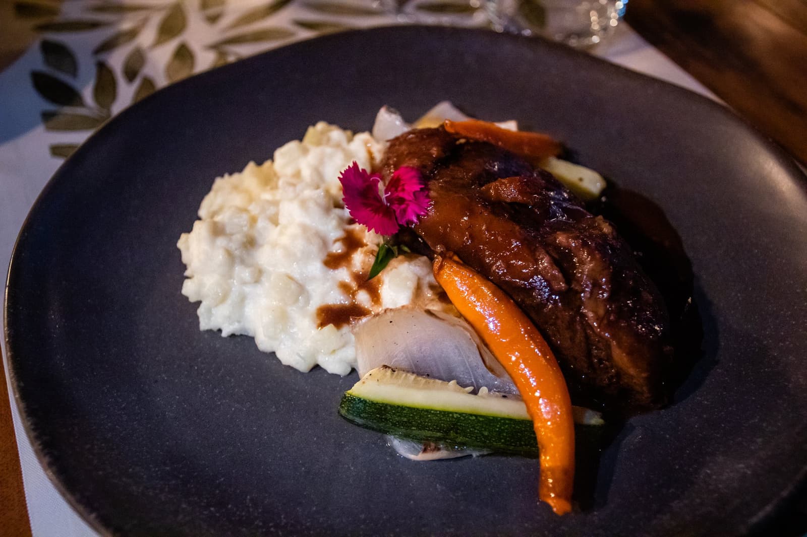 Dinner at the estancia: slow-braised, flower-topped, and worth every cold kilometer getting here. A dark ceramic plate at Estancia Cerro Guido restaurant, featuring a braised meat portion glazed in rich dark sauce, served alongside creamy risotto and roasted vegetables including carrot and zucchini, garnished with a small pink edible flower