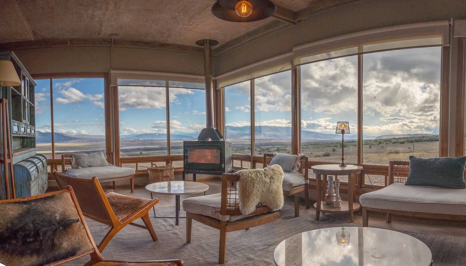 Fire going, steppe rolling out to the horizon, nowhere you need to be. Panoramic lounge at Estancia Cerro Guido with a lit wood-burning stove, leather and sheepskin chairs, and floor-to-ceiling windows framing a wide view of the Patagonian steppe and distant mountains under a partly cloudy sky