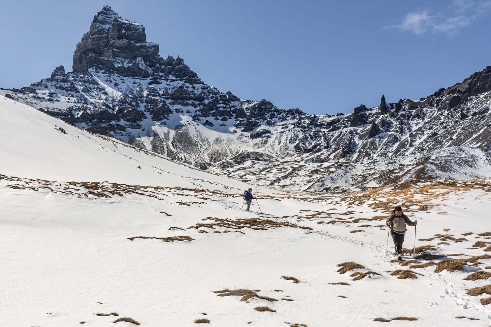 Fourteen kilometres, a frozen valley, and that rock tower daring you to keep going. Photo: Gonzalo Vargas Two hikers with trekking poles ascend a snow-covered slope at estancia cerro guido, with jagged dark rock towers rising above a broad white valley under a clear blue sky