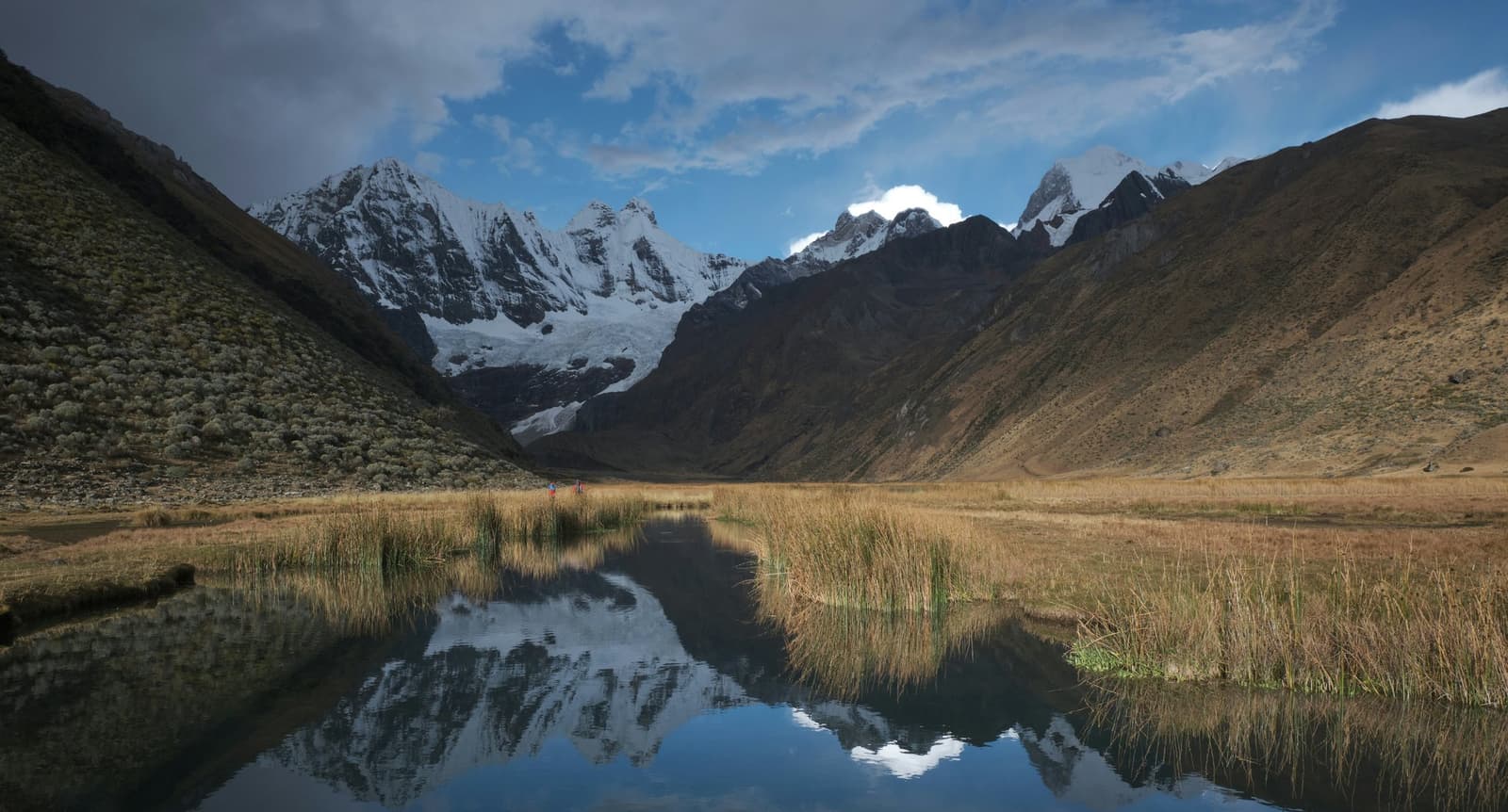 The mountains don't care how small you are, the pond will remind you anyway. A still alpine pond on the huayhuash circuit reflects a row of jagged, snow-capped peaks rising above a broad high-altitude valley, with golden puna grassland and reed clumps in the foreground and two tiny trekkers visible in the middle distance
