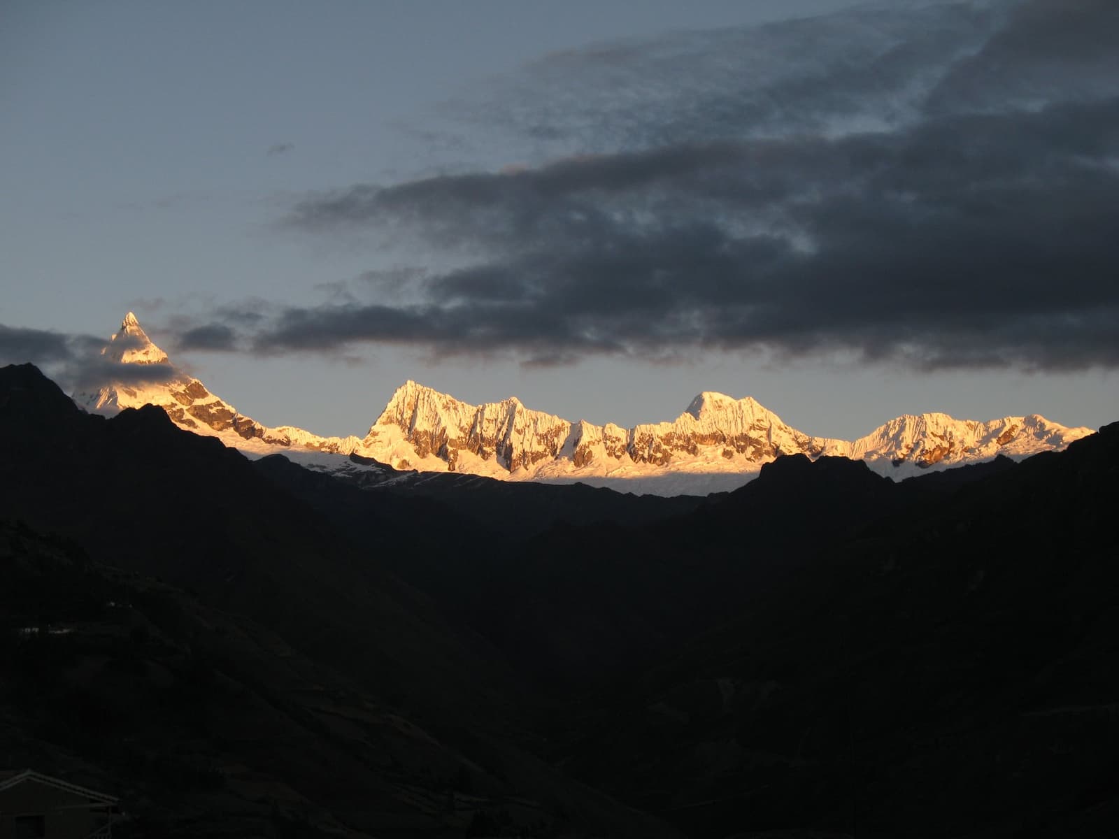 That window between cloud and ridge, lit up just long enough to matter. Snow-capped peaks of the cordillera huayhuash glowing amber and gold under the last light of sunset, with dark ridge silhouettes in the foreground and a band of heavy cloud above